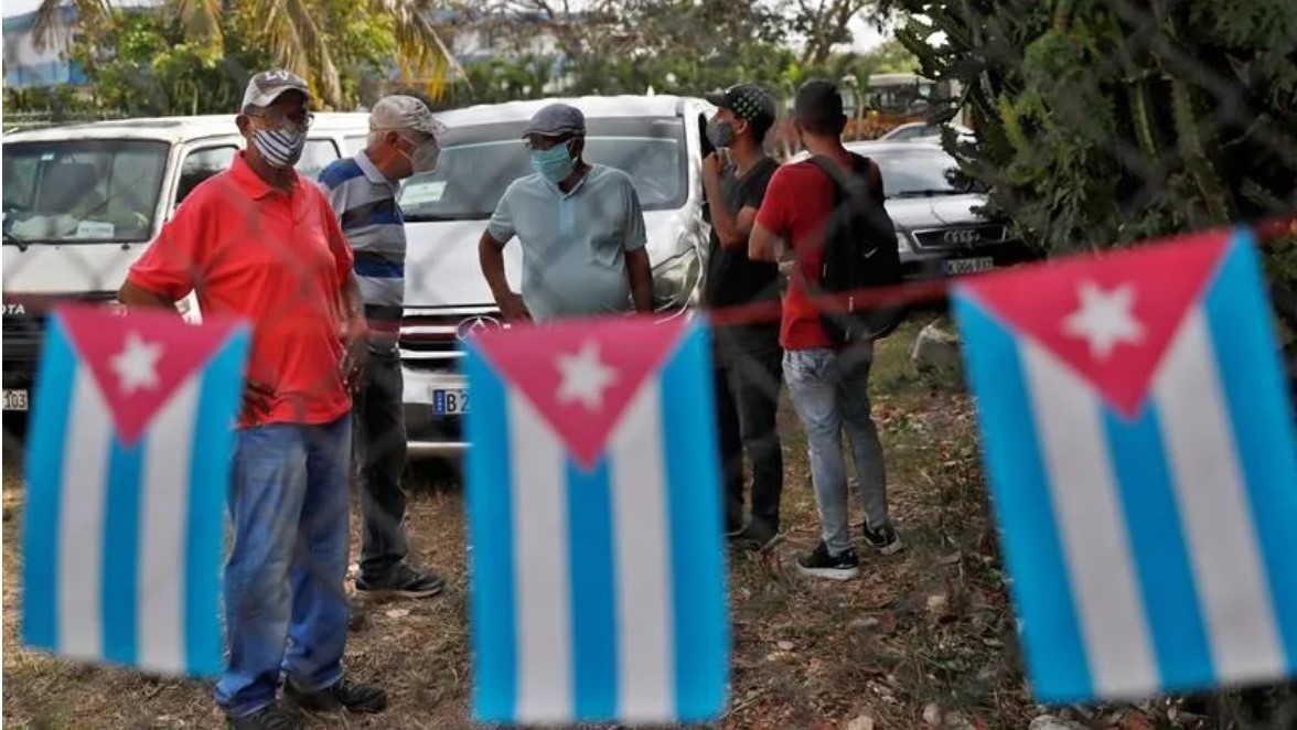Cubanos con mascarillas tras una cerca con banderitas cubanas.