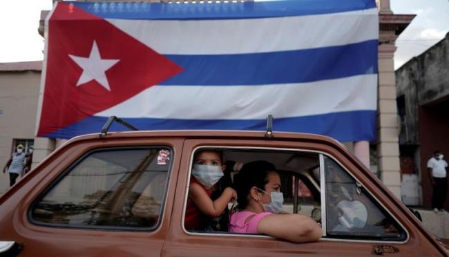 Una familia en un auto pasando por delante de una bandera de Cuba en La Habana.