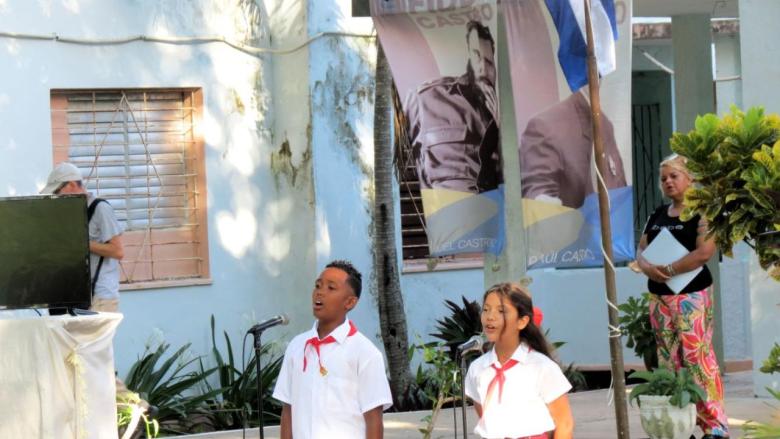 Dos niños de primaria en un acto escolar con imágenes de Raúl y Fidel Castro detrás.